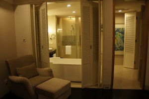 The image shows a cozy, well-lit bathroom and adjacent seating area. The bathroom features a modern design with a glass-enclosed shower, a white bathtub, and a towel hanging on the tub. There is a vanity with a mirror and a small round mirror mounted on the wall. The seating area includes a comfortable beige armchair with a matching ottoman. The room is separated by white folding doors, and there is a piece of artwork visible on the wall in the background.