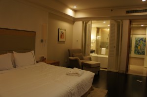 The image shows a well-lit hotel room with a large bed on the left side, covered in white linens. There is a bedside table next to the bed with a lamp and a small item on it. An armchair with an ottoman is positioned near the foot of the bed. In the background, there is an open doorway leading to a bathroom, which is also well-lit. The bathroom has a bathtub and a large mirror. The walls are decorated with framed artwork. The overall ambiance of the room is cozy and inviting.