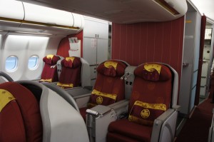 The image shows the interior of an airplane's business class cabin. The seats are wide and spacious, upholstered in red fabric with yellow accents and decorative patterns. Each seat has a headrest and a pillow. The cabin has a clean and modern design, with windows on the left side and a partition wall towards the front. The overall atmosphere appears comfortable and luxurious.
