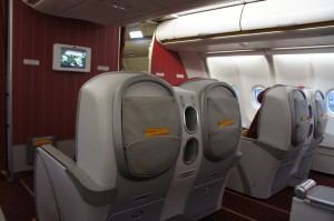 The image shows the interior of an airplane's business or first-class cabin. The seats are large and equipped with personal entertainment screens. The seats are arranged in a spacious layout, with ample legroom and privacy partitions. The cabin has a modern design with red and gray color tones. The windows are visible on the right side of the image, and the overhead compartments are closed.