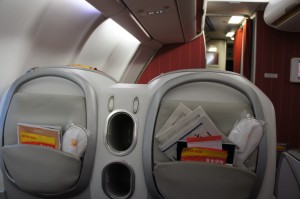 The image shows the back of two airplane seats in a cabin. Each seat has a pocket containing various items such as safety information cards, magazines, and possibly a pair of headphones or a small pillow. The seats are separated by a panel with two circular openings, likely for holding drinks. The cabin interior is visible in the background, with overhead compartments and a red curtain.