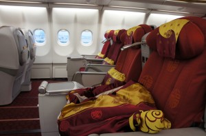 The image shows the interior of an airplane cabin, specifically the business or first-class section. The seats are upholstered in red fabric with yellow accents and patterns. Each seat has a headrest cover and a blanket draped over it. The cabin has large windows, and the seats are arranged in a spacious layout with ample legroom. The overall setting appears clean and comfortable.