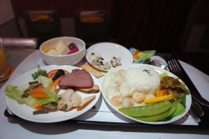 The image shows a meal served on a tray, likely on an airplane. The tray contains several dishes, including a plate with rice and shrimp in a creamy sauce, a side of vegetables, and another plate with assorted items such as sliced meat, mushrooms, and more vegetables. There is also a small bowl of fruit and a small dish with what appears to be a dessert or snack. Utensils are placed on the tray as well.