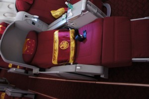 The image shows a luxurious airplane seat in a business or first-class cabin. The seat is upholstered in red fabric and is equipped with a headrest and a footrest. There is a yellow and red blanket with a logo draped over the seat. The seat has a personal entertainment screen, a small table with a water bottle, and other amenities. The surrounding area includes other similar seats, indicating a spacious and comfortable seating arrangement.