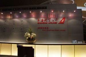 The image shows the reception area of the Shanghai Airlines First Class Lounge. The wall behind the reception desk features the Shanghai Airlines logo and name in both English and Chinese. There is a large flower arrangement on the counter, and the area is well-lit with overhead lights. The design is modern and sleek, with a combination of metallic and warm tones.