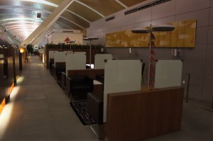 The image shows a modern, well-lit airport lounge with individual seating areas. Each seating area is separated by partitions for privacy and includes a chair and a small table. The lounge has a high, curved ceiling with a series of lights along the top. There are decorative elements on the walls, including a large, abstract painting. The overall atmosphere appears to be quiet and comfortable, suitable for travelers to relax or work while waiting for their flights.