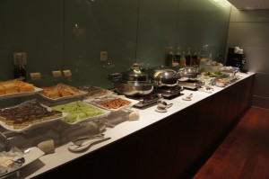 The image shows a buffet setup with various food items displayed on a long counter. There are several trays containing different dishes, including what appears to be salads, desserts, and hot foods in chafing dishes. There are also plates, utensils, and condiments available for guests to use. The background features a glass wall, and the counter is well-lit, creating an inviting atmosphere for dining.