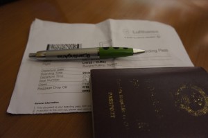 The image shows a boarding pass, a pen, and a passport placed on a wooden surface. The boarding pass is partially visible, with some text and a barcode. The pen is placed on top of the boarding pass, and the passport is partially covering the bottom right corner of the boarding pass. The passport has a dark cover with gold text and emblem.