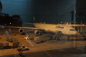 The image shows a Lufthansa airplane parked at an airport gate at night. The aircraft is being serviced, with various ground support equipment and vehicles around it. The scene is dimly lit, and the reflection of the terminal windows is visible in the foreground.