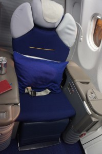 The image shows a business or first-class airplane seat. The seat is upholstered in dark blue fabric and has a matching blue pillow placed on it. There is a seatbelt on the seat. The surrounding area includes a small table with a can of soda and a red notebook on it. The seat is positioned next to a window, and the window shade is partially open. The armrests and side panels are in a light beige color.