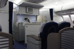 The image shows the interior of an airplane cabin, specifically the business or first-class section. There are several large, cushioned seats with headrests, arranged in rows. In the background, there is a galley area with a countertop, cabinets, and a microwave. The cabin is well-lit, and there are curtains that can be drawn for privacy. The overall atmosphere appears clean and organized.