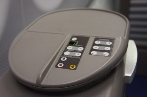 The image shows a close-up of an airplane seat control panel. The panel is integrated into the armrest and features various buttons for adjusting the seat position. The buttons include icons for reclining the seat, adjusting the leg rest, and other seat functions. The panel is gray with a combination of black and white buttons, and some buttons have colored indicators.