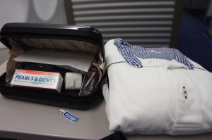 The image shows a small black toiletry bag that is open, revealing its contents, which include a box of Pearls & Dents toothpaste, a toothbrush, and other personal care items. Next to the toiletry bag is a neatly folded white shirt with a blue and white checkered collar. There is also a small sticker or label on the surface in front of the toiletry bag. The items are placed on a flat surface, possibly a table or countertop.