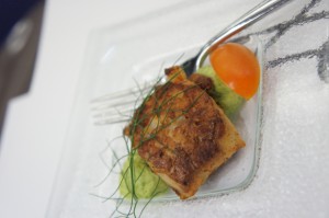 The image shows a plated dish consisting of a piece of cooked fish garnished with herbs, placed on a bed of green puree, and accompanied by a small orange vegetable, possibly a cherry tomato. A fork is placed next to the food on the plate.