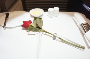 The image shows a table set for a meal with a white tablecloth. On the table, there is a single red rose in a small glass vase, a plate with a knife, a small dish with a pat of butter, and two small white containers, likely for salt and pepper. There is also a neatly rolled white napkin with a silver napkin ring.