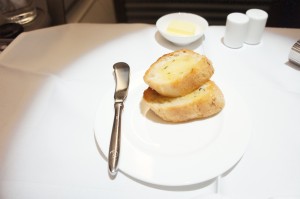 The image shows a white plate with two pieces of garlic bread on it. A butter knife is placed on the edge of the plate. In the background, there is a small dish with a pat of butter and two small white containers, likely for salt and pepper. The setting appears to be on a white tablecloth.