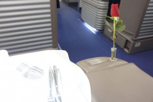 The image shows a close-up of a dining setup in what appears to be a luxurious setting, possibly an airplane's first-class cabin. There is a white tablecloth with neatly arranged silverware, including forks and knives. To the right, there is a single red rose in a small vase attached to a beige surface. The background features blue carpeted flooring and partitioned sections, suggesting a high-end travel environment.