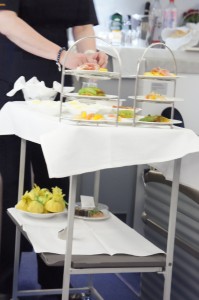 A person is standing next to a serving cart covered with a white tablecloth. The cart has multiple tiers with various plated food items, including what appears to be small sandwiches, fruits, and pastries. There are also some wrapped items on a lower shelf of the cart. The background shows a countertop with various bottles and containers.
