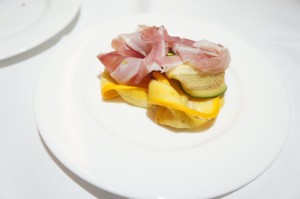 A white plate with a small serving of food, consisting of thinly sliced cured meat, possibly prosciutto, layered over slices of yellow and green vegetables, possibly squash and zucchini. The plate is set on a white tablecloth.