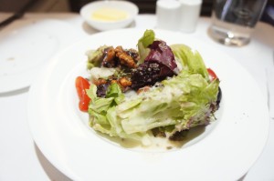 A white plate with a fresh salad consisting of various types of lettuce, cherry tomatoes, and what appears to be nuts and possibly a creamy dressing. In the background, there is a small dish with a yellow substance, likely butter, and a glass of water.
