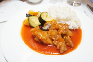 A plate of food featuring a serving of white rice, pieces of chicken in a red sauce, and a side of mixed vegetables including zucchini and yellow squash. The dish is presented on a white plate.