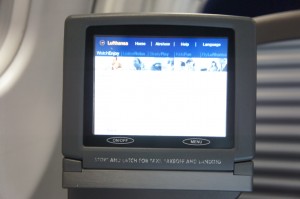 A close-up image of an in-flight entertainment screen on an airplane. The screen displays the Lufthansa logo and menu options such as "Home," "Airline," "Help," and "Language." The screen is mounted on the back of an airplane seat, and the background shows part of the airplane's interior.