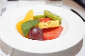 A white bowl containing a variety of fresh fruit slices, including orange segments, kiwi slices, a piece of pineapple, a red grape, and a slice of watermelon. There is also a sprig of mint on top of the fruit. The bowl is placed on a white surface with a knife partially visible in the background.