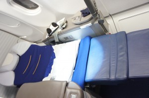 The image shows a fully reclined airplane seat in a business or first-class cabin, forming a flat bed. The seat is upholstered in blue and gray fabric, with a white blanket or sheet covering part of it. The surrounding area includes a window, a control panel, and a small storage compartment.