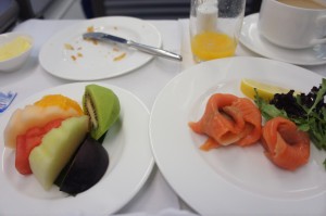 The image shows a meal served on a white tablecloth. There are two plates: one with assorted fresh fruit slices including watermelon, kiwi, and melon, and another with smoked salmon, a lemon wedge, and a small salad. In the background, there is a glass of orange juice, a cup of coffee, and a small bowl of butter. A knife and a plate with some crumbs are also visible.