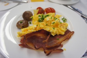 A white plate with a breakfast meal consisting of scrambled eggs garnished with chopped herbs, crispy bacon strips, two small whole mushrooms, and two cherry tomatoes. The plate is set on a table with a fork and knife visible in the background.