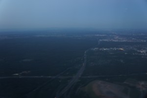 The image shows an aerial view of a landscape during dusk or dawn. The scene includes a vast expanse of land with a mix of forested areas and open fields. A road or highway runs through the middle of the image, leading towards a distant city or town with visible buildings and lights. The sky above is clear with a gradient from light to dark blue.