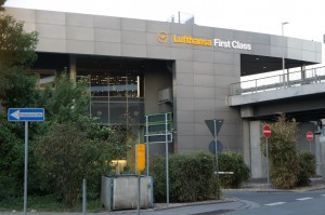 The image shows the exterior of a building with a sign that reads "Lufthansa First Class." The building has a modern design with large windows and is surrounded by greenery. There are several road signs and a pedestrian walkway in the foreground. A bridge or elevated roadway is visible on the right side of the image.