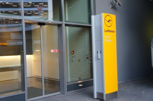 The image shows the entrance to a Lufthansa First Class lounge. The entrance features sliding glass doors and a prominent yellow sign with the Lufthansa logo and the words "Lufthansa First Class" written on it. The area appears clean and modern, with a glimpse of the interior visible through the glass doors.