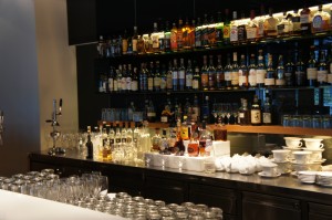 A well-stocked bar with a variety of liquor bottles arranged on shelves behind the counter. The counter has several rows of clean glasses, a few coffee cups stacked on saucers, and some bar equipment. The lighting is warm, creating a cozy atmosphere.