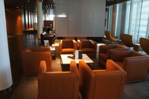 The image shows a modern, well-lit lounge area with several brown leather armchairs arranged around small square tables. The space has a contemporary design with large windows allowing natural light to enter. The floor is dark, and there are white columns and a large white wall in the background. The overall atmosphere appears to be comfortable and inviting, suitable for relaxation or casual meetings.