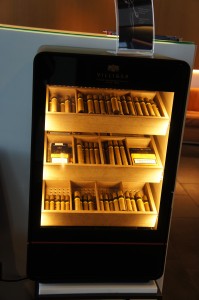 The image shows a well-lit humidor cabinet filled with various cigars. The cigars are neatly arranged on multiple shelves inside the cabinet. The humidor has a glass door, allowing the cigars to be visible. The interior lighting highlights the different sizes and types of cigars stored within.