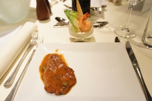 A white plate with a small portion of meat covered in a brown sauce is placed on a table set for a meal. In the background, there is a small glass containing a shrimp and some greens. The table is elegantly set with silverware, a wine glass, and a white napkin.