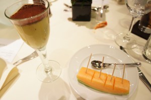 The image shows a dessert and a fruit plate on a table. The dessert is served in a tall glass and appears to be a layered pudding or mousse with a dusting of cocoa powder on top. Next to it, on a white plate, there are several slices of cantaloupe with toothpicks inserted into them, along with a small spoon. The table is set with cutlery, glasses, and other dining items in the background.