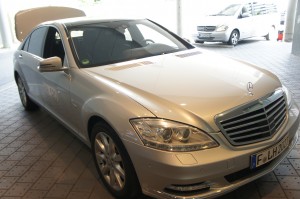 A silver Mercedes-Benz sedan is parked indoors with its trunk open. Another vehicle is visible in the background. The car has a European license plate.
