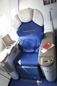 The image shows a luxurious airplane seat in a business or first-class cabin. The seat is upholstered in blue and gray, with a large blue pillow placed on it. There is a small table attached to the side of the seat, which holds a red passport and a boarding pass. The seat has various controls and compartments, indicating it is designed for comfort and convenience during a flight. The window of the airplane is visible in the background.