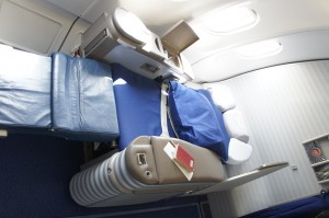 The image shows a first-class or business-class airplane seat that has been fully reclined into a flat bed. The seat is covered with a blue blanket and has a pillow at the headrest. There is a small table next to the seat with a closed laptop and a red book on it. The area is well-lit with natural light coming from the airplane windows. The overall setting appears to be comfortable and designed for long-haul flights.