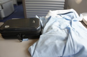 A small black suitcase and a light blue shirt are placed on a table. The background shows a part of a room with metallic surfaces and a blue floor.
