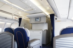The image shows the interior of an airplane's first-class or business-class cabin. The seats are spacious and upholstered in blue fabric with white headrests. There is a partition wall with a small screen mounted on it, and overhead storage compartments are visible. The cabin has a clean and modern design, with soft lighting and a curtain separating different sections.