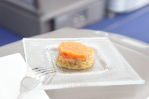 A small piece of food, possibly a dessert or appetizer, is placed on a square glass plate. The food item has a layered appearance with an orange top layer and a yellowish bottom layer. A fork is placed next to the plate. The background is slightly blurred, suggesting the image was taken in a setting like a restaurant or an airplane.