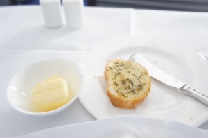A slice of bread with herbs on a white plate, accompanied by a butter knife. Next to the plate is a small bowl containing a piece of butter. The background includes a white tablecloth and two white containers, likely for salt and pepper.