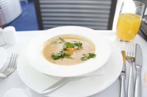 A bowl of soup garnished with herbs is placed on a white plate with a napkin and a spoon beside it. There is a glass of orange juice to the right of the plate, and a fork and knife are also visible on the right side of the image. The background shows a blurred setting, possibly a dining area.