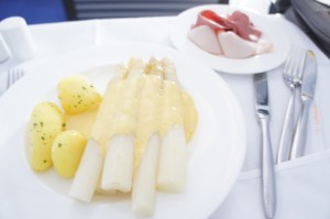 The image shows a plate of food consisting of white asparagus topped with a creamy yellow sauce, accompanied by boiled potatoes garnished with herbs. In the background, there is a small plate with slices of ham. The table is set with a knife, fork, and spoon.