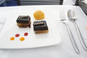 A white plate with two small, square chocolate desserts, each topped with a decorative piece, and a scoop of orange sorbet. There are also three small dots of red and orange sauce on the plate. Next to the plate, there is a fork, a spoon, and a knife placed on a white tablecloth.