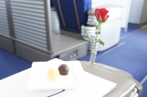 The image shows a white plate with two pieces of chocolate, one white and one dark, placed on a surface. Next to the plate, there is a single red rose in a small vase. The background appears to be the interior of an airplane, with blue seats and a modern design.