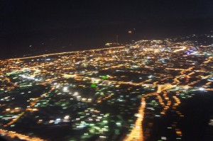 Aerial view of a city at night, with numerous lights illuminating the streets and buildings. The lights create a network of glowing lines and clusters, giving a sense of the city's layout and activity. The image is slightly blurred, adding a sense of motion.